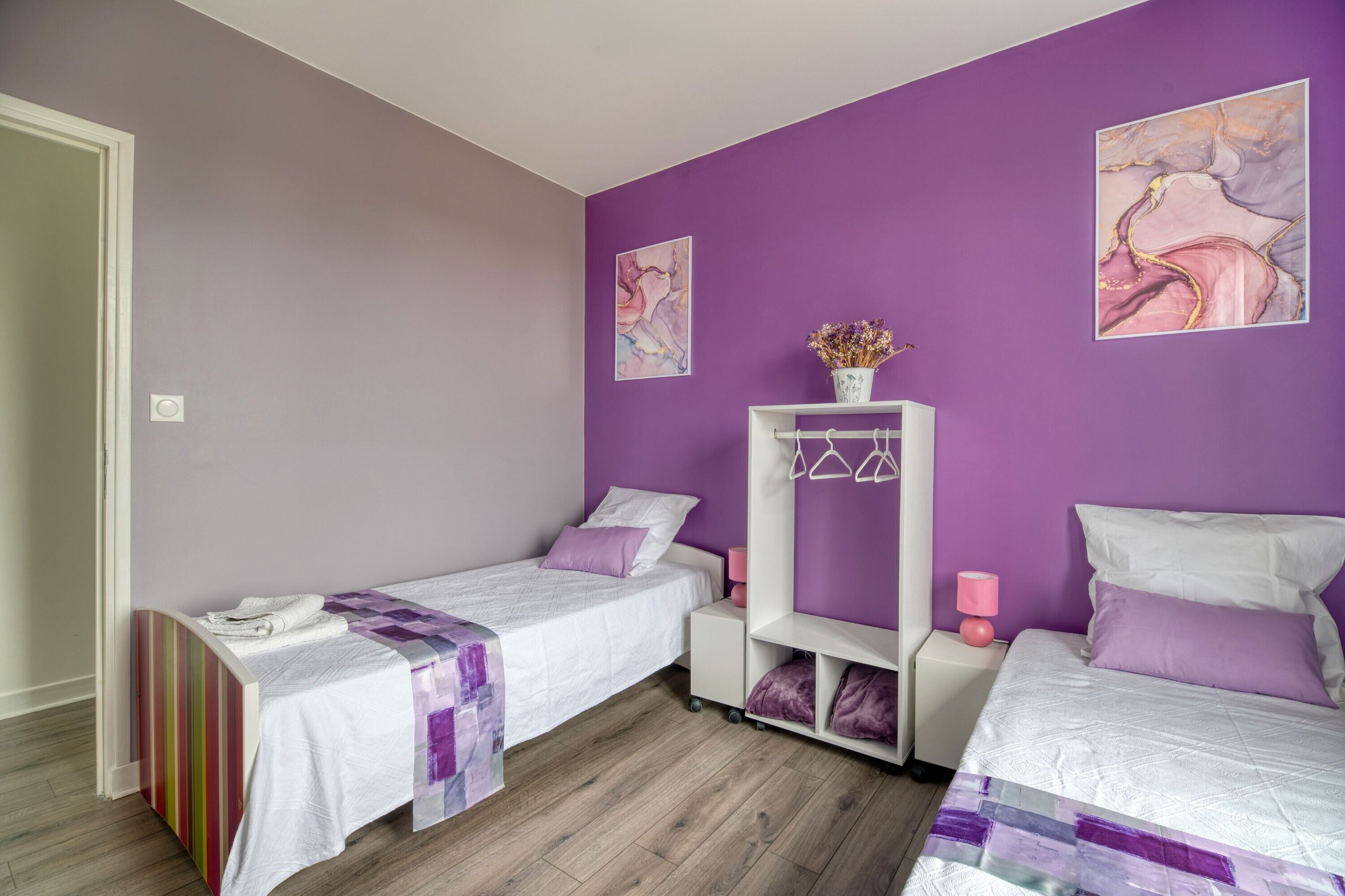 Une chambre avec deux lits, une table de chevet blanche, une commode blanche, des murs violets et des œuvres d’art.