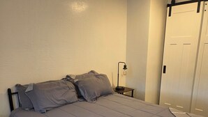 A bed with a grey duvet, a bedside lamp, a wooden nightstand, and a white door. - Country Railroad - Wyoming Primo Managment (Green River)
