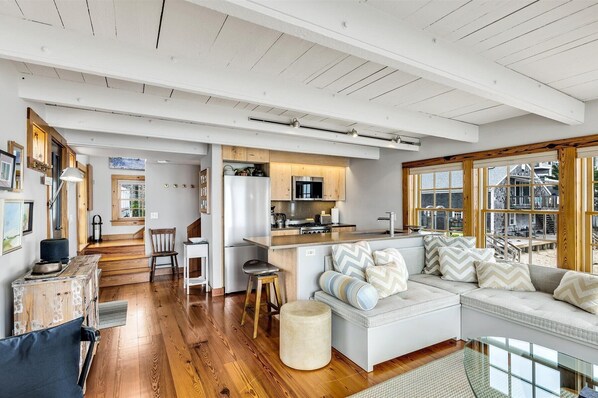 A spacious living area with a wooden floor, a white sofa with patterned cushions, a kitchen with wooden cabinets, and large windows offering a view of the outside.