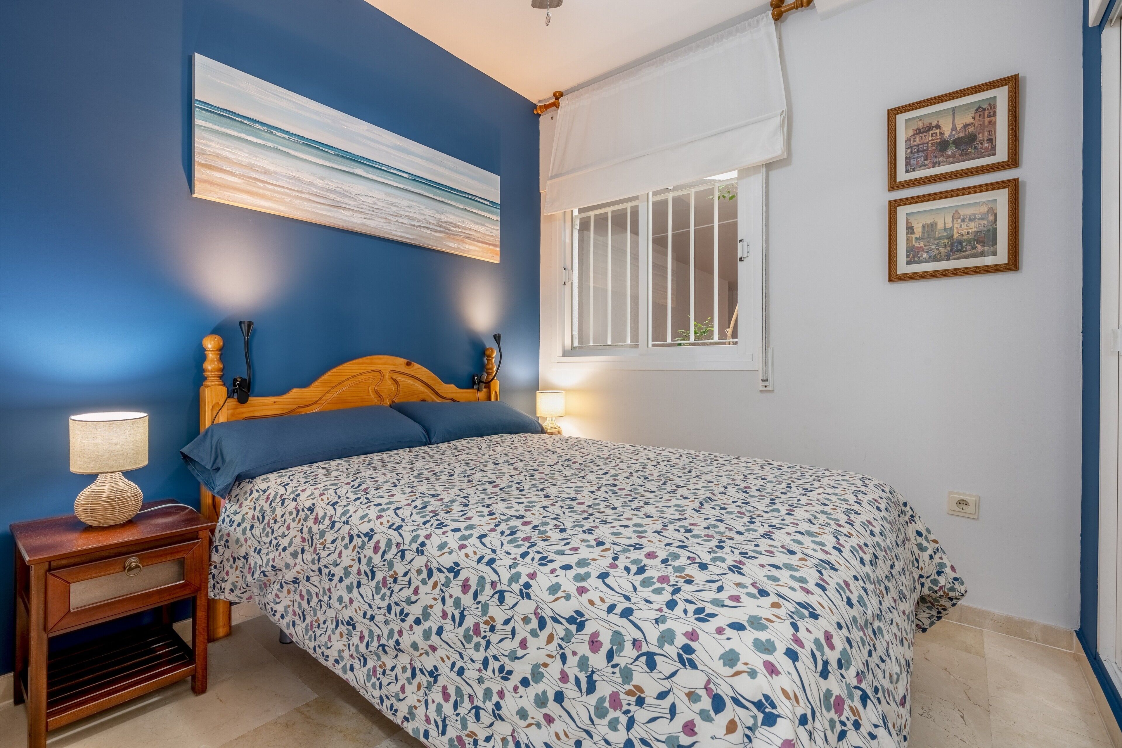 A bedroom with a bed, a nightstand, a lamp, a window with white curtains, and two framed pictures on the wall.