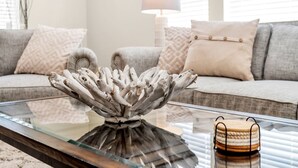 A living room with a glass coffee table, a gray sofa with patterned cushions, and a lamp in the background.