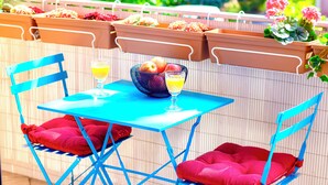 Et blåt bord med en skål frugt og et glas appelsinjuice, to blå stole med røde hynder, og terracotta krukker med blomster og grøntsager på en balkon.