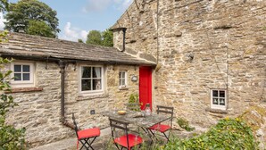 A stone cottage with a red door, a small window, and a table with red chairs.