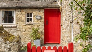 A stone cottage with a red door, a small window with curtains, and a red picket fence.