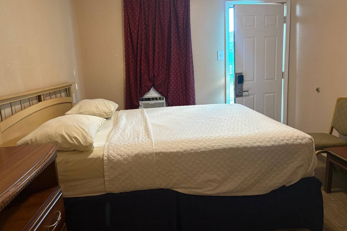 Une chambre d’hôtel avec un lit, une table de chevet en bois, une chaise, une fenêtre avec des rideaux rouges et une porte.