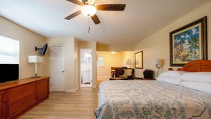 Une chambre à coucher avec un lit, un ventilateur de plafond, un téléviseur posé sur une commode en bois et une porte donnant sur une autre pièce.