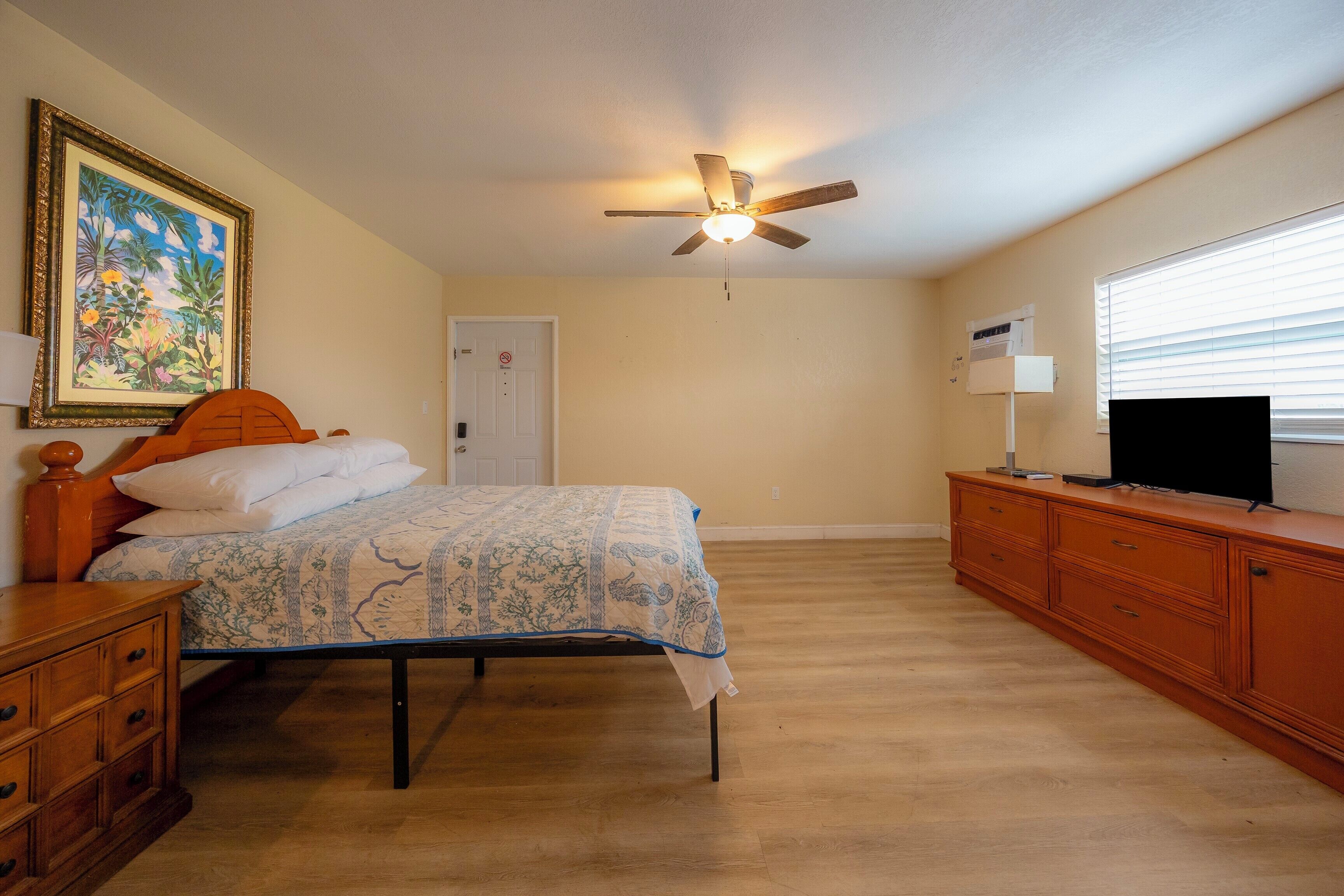 A bedroom with a bed, dresser, ceiling fan, and a painting on the wall.