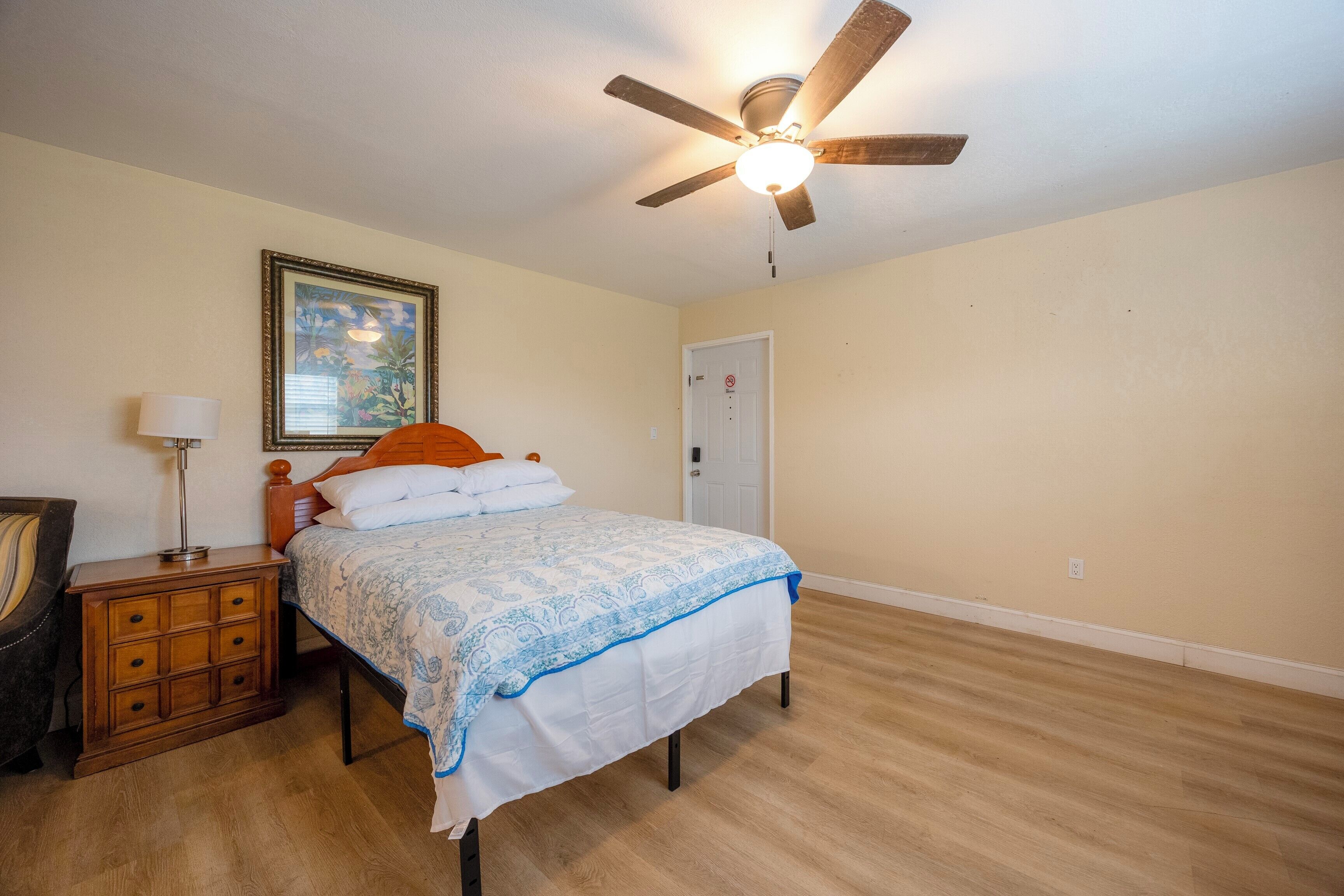 A bedroom with a bed, a nightstand, a lamp, a ceiling fan, a door, and a framed picture on the wall.