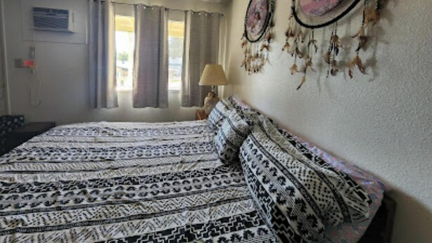 A bedroom with a patterned bedspread, curtains, a wall-mounted dreamcatcher, and a lamp. - Columbia Inn Motel CA 49 (Columbia)