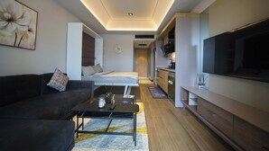 A modern living room with a sofa, coffee table, and a TV. There is a bedroom with a bed and a bathroom visible in the background.