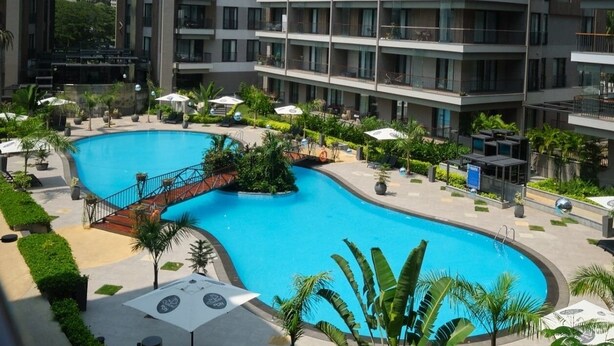 A modern hotel with a large swimming pool, multiple balconies, and a view of a power plant in the background.