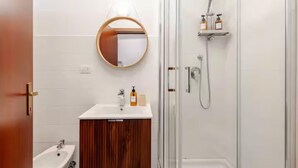 Een moderne badkamer met een houten wastafelmeubel, een ronde spiegel, een douche met een handdouche en een klein plankje met toiletartikelen.