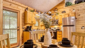 A wooden cabin-style kitchen with wooden cabinets, a stainless steel refrigerator, and a dining table set with black bowls and a white pitcher. - Er80 - Can't Bear To Leave 2 Bedroom Cabin (Pigeon Forge)