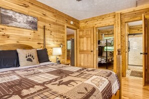 Quarto de cabana de madeira com cama, mesa de cabeceira, abajur e porta para outro cômodo.