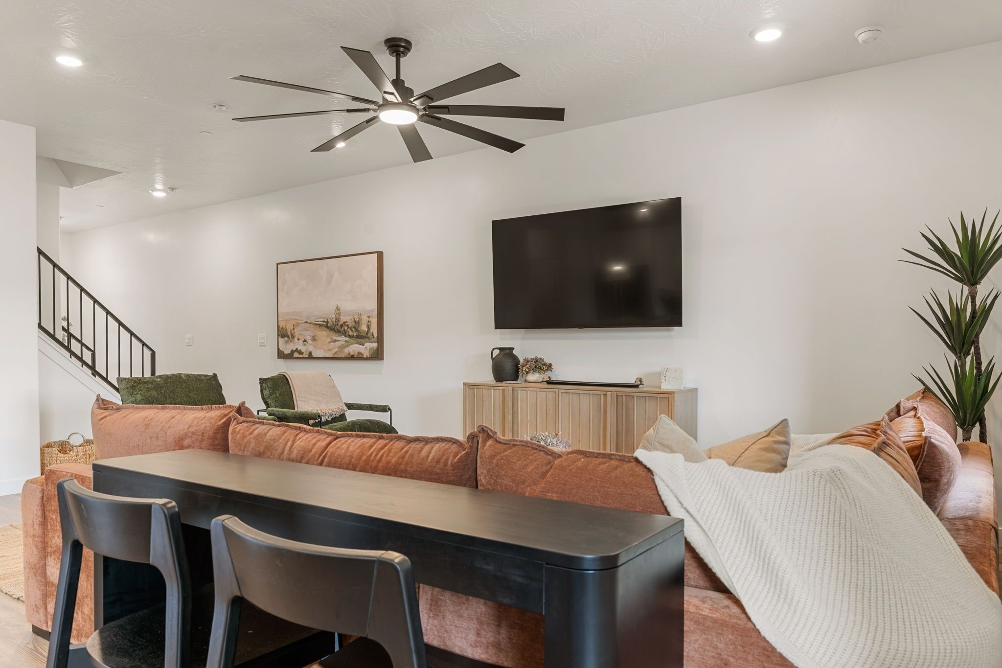 A modern living room with a dining area, a flat-screen TV mounted on the wall, a ceiling fan, and a staircase leading to another level.