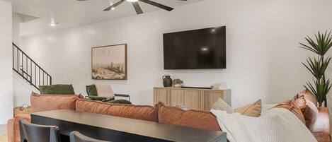 A modern living room with a dining area, a flat-screen TV mounted on the wall, a ceiling fan, and a staircase leading to another level.