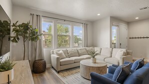 A modern living room with a beige sofa, a blue armchair, and a round coffee table. There are large windows with curtains, a flat-screen TV mounted on the wall, and a plant on a wooden table. - Relax In Style 4 Bedroom Townhouse (Washington)