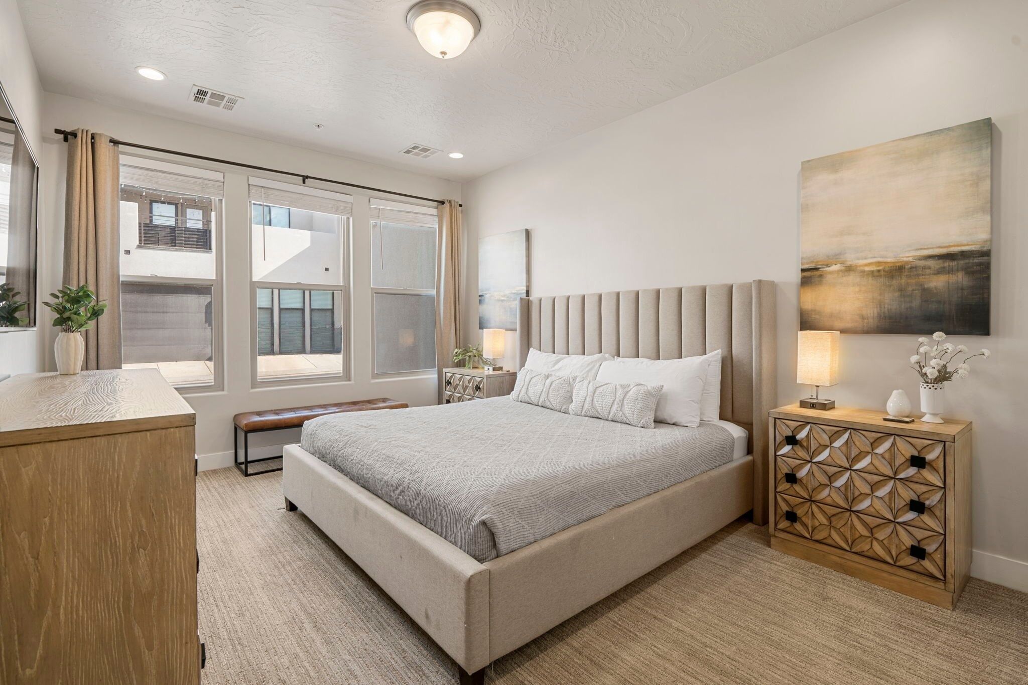 A modern bedroom with a large bed, a wooden nightstand, a bench, and a view of the outside.