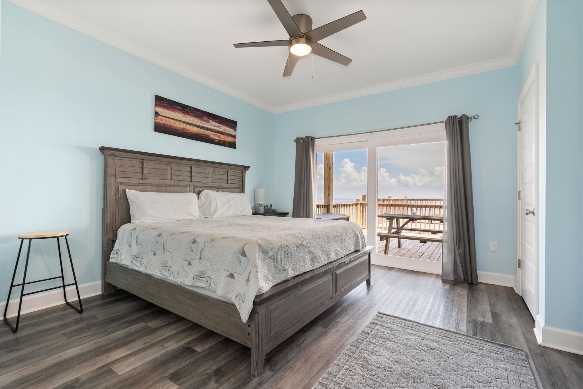 A bedroom with a bed, a ceiling fan, a nightstand, a window with curtains, and a view of an outdoor deck.