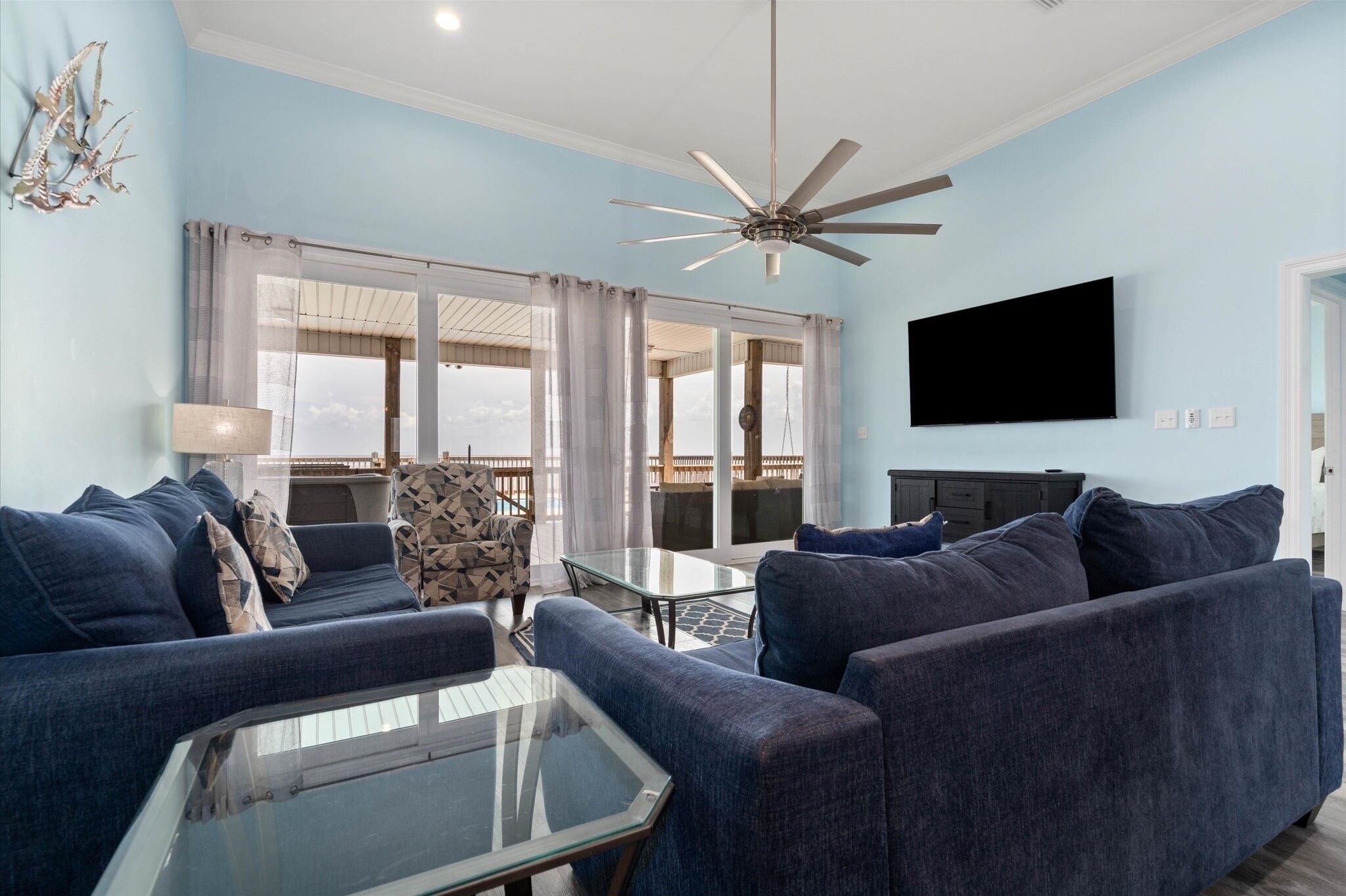 A living room with a ceiling fan, a flat-screen TV, and a view of the outdoors through large windows.