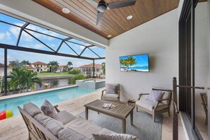 A modern outdoor lounge area with a pool, comfortable seating, and a large TV screen displaying a tropical beach scene.