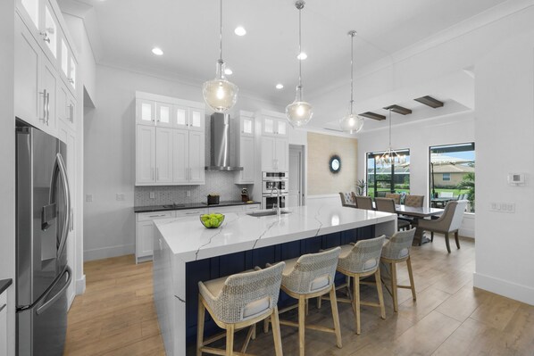 A modern kitchen with white cabinets, a stainless steel refrigerator, a large island with bar stools, and pendant lighting.