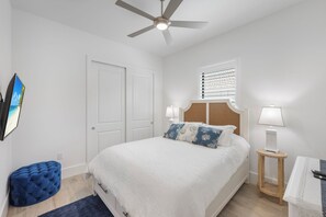 A bedroom with a bed, a ceiling fan, a window with blinds, a bedside table with a lamp, and a blue ottoman.