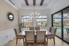 A dining area with a glass table and four upholstered chairs, a chandelier, and a view of a swimming pool and outdoor patio.