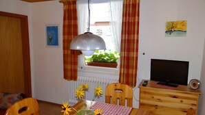 Een kamer met een eettafel, stoelen, een televisie, een radiator, een raam met gordijnen en een kamerplant.