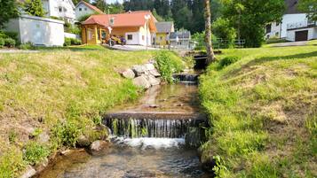 Ein kleiner Bach mit einem Wasserfall, umgeben von grünen Ufern und Wohnhäusern im Hintergrund.