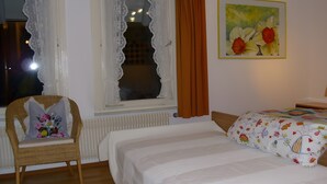 Un dormitorio con cama, una silla con cojín, una ventana con cortinas y un cuadro en la pared.