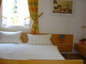 A bed with white bedding, a wooden headboard, a nightstand with a lamp, a window with curtains, and a wall-mounted picture.