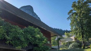 Un edificio de madera tradicional con techo inclinado, rodeado de abundante vegetación y montañas al fondo.