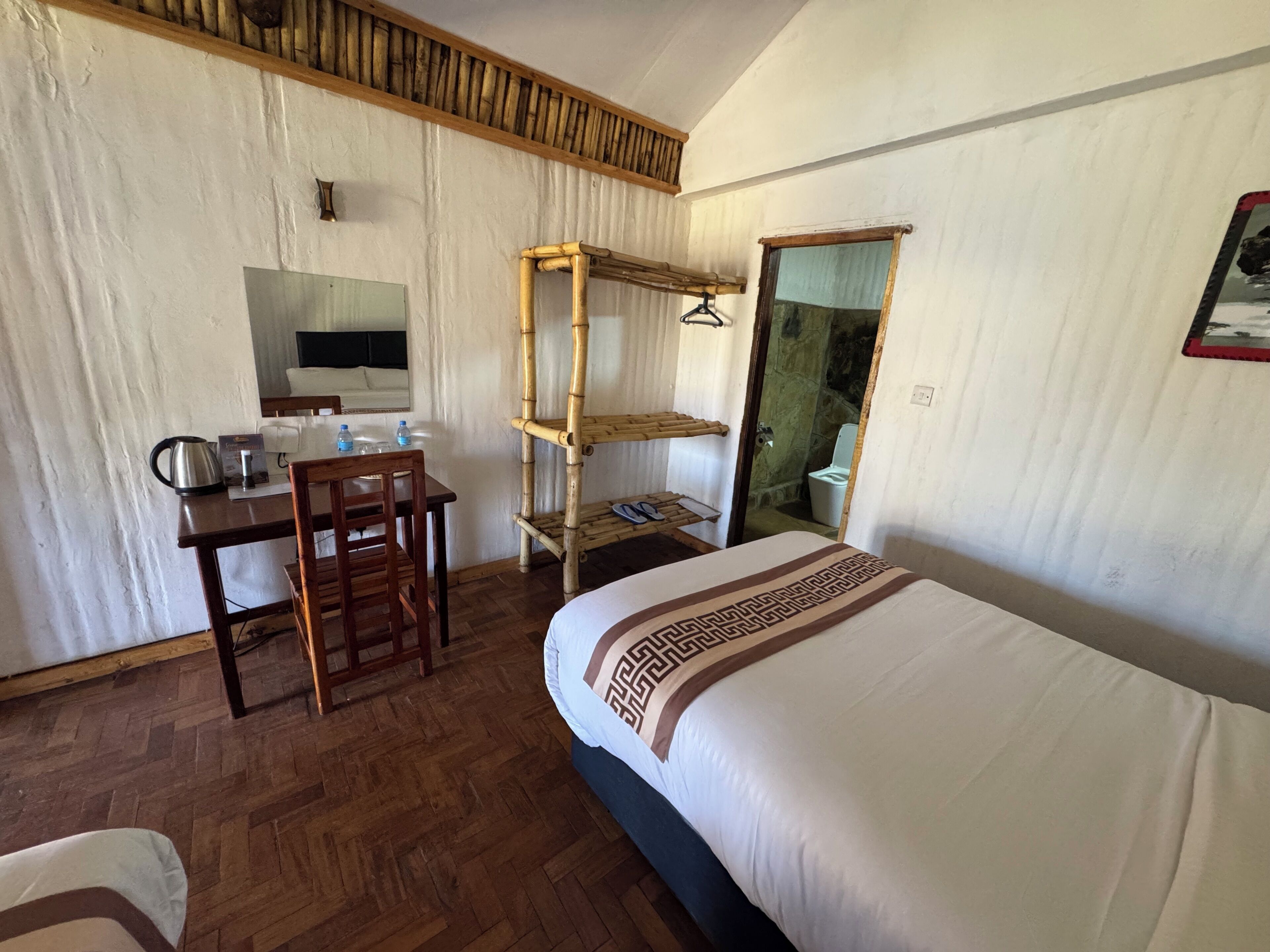 A hotel room with a bed, a desk with a chair, a wooden shelf, and a bathroom visible through an open door.