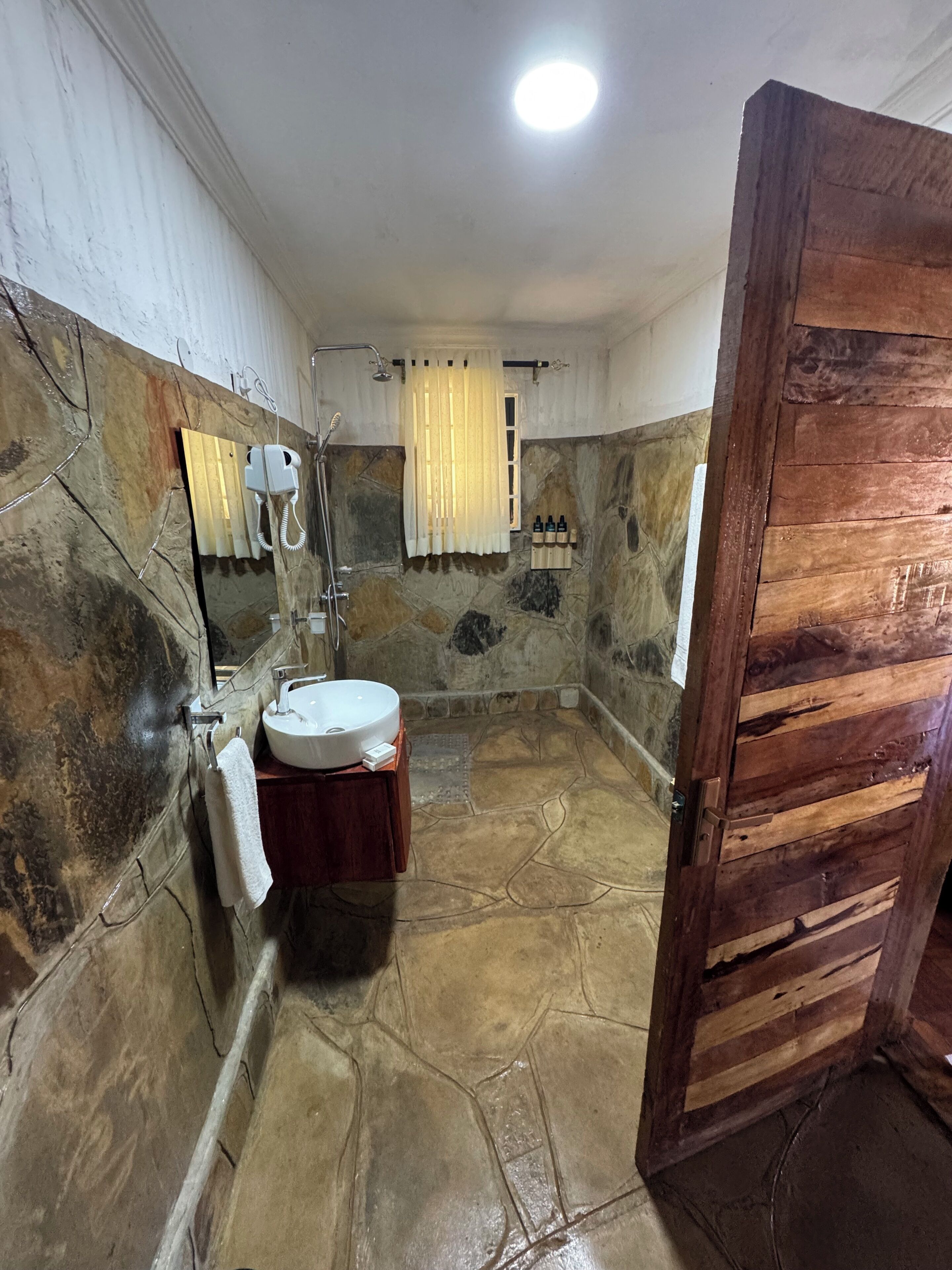 Un baño con pared de piedra, puerta de madera, lavamanos blanco y zona de ducha.