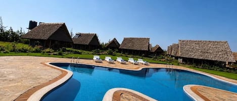 A swimming pool with a unique wavy design, surrounded by thatched-roof cottages and lounge chairs.