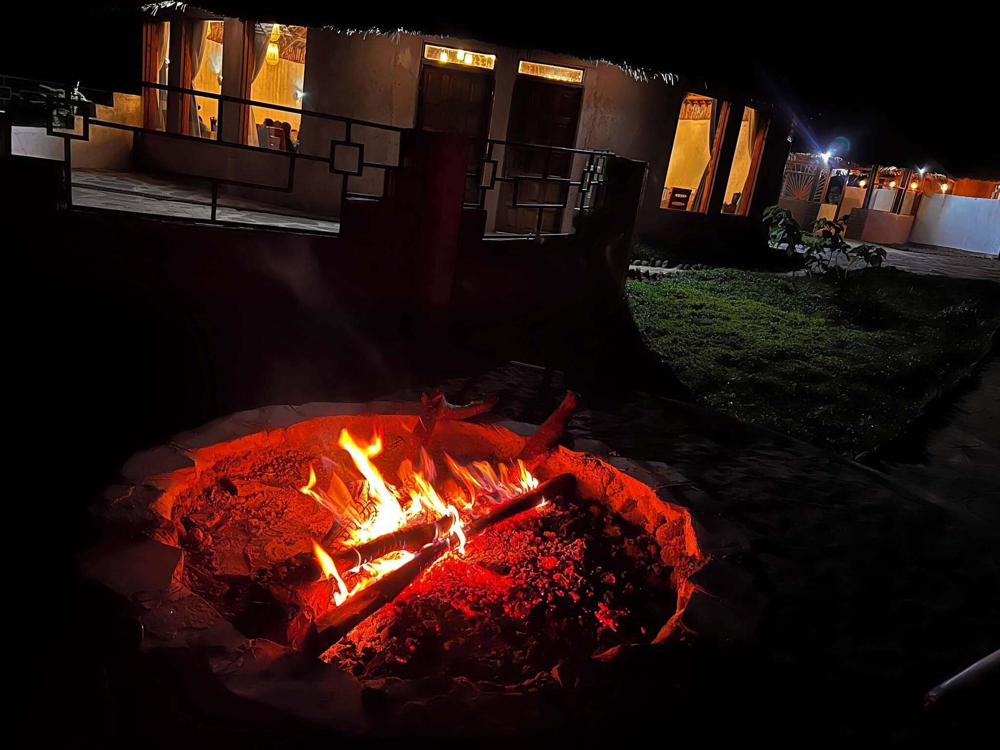 A nighttime outdoor setting with a fire pit in the foreground and a building with illuminated windows in the background.