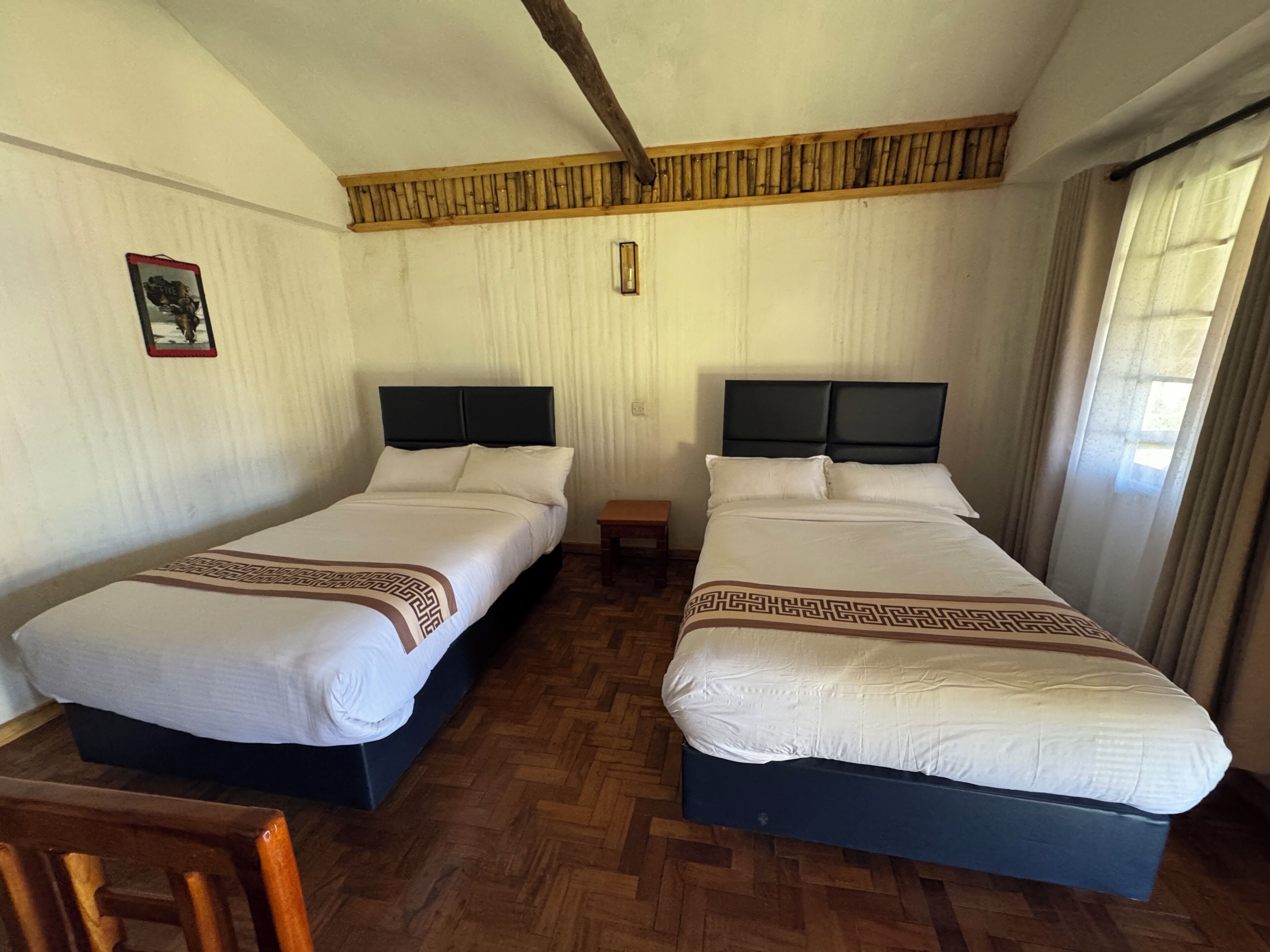 Una habitación con dos camas, suelo de madera, viga de techo de madera, una mesita y un cuadro en la pared.