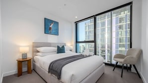 Un dormitorio moderno con una cama grande, una mesita de noche, una silla y vistas a la ciudad a través de ventanales de gran altura.