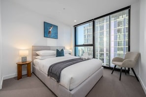 A modern bedroom with a large bed, a side table, a chair, and a city view through floor-to-ceiling windows.