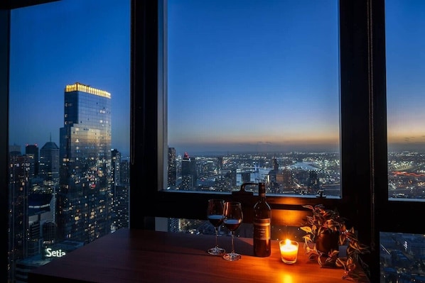 A cityscape view from a window with a table featuring a bottle, glasses, and a candle.