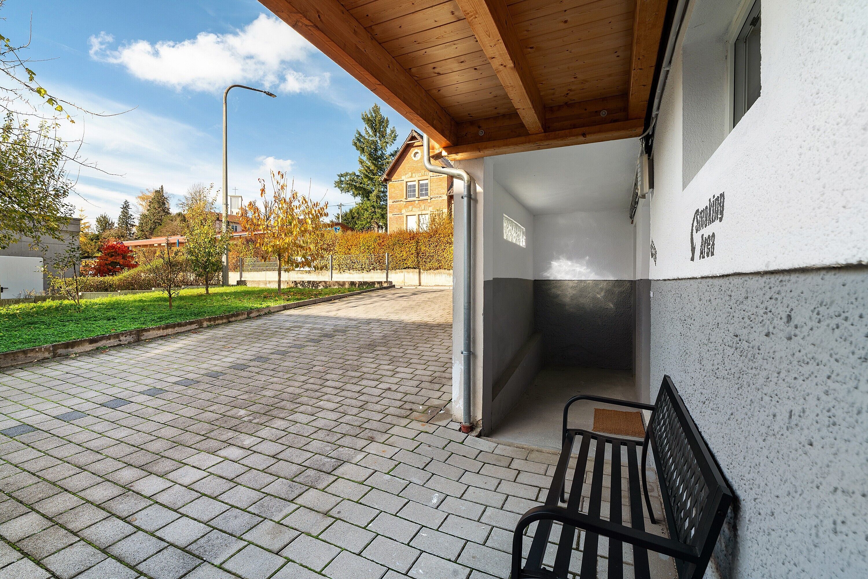 A covered outdoor area with a bench, a paved walkway, and a building with a sign.