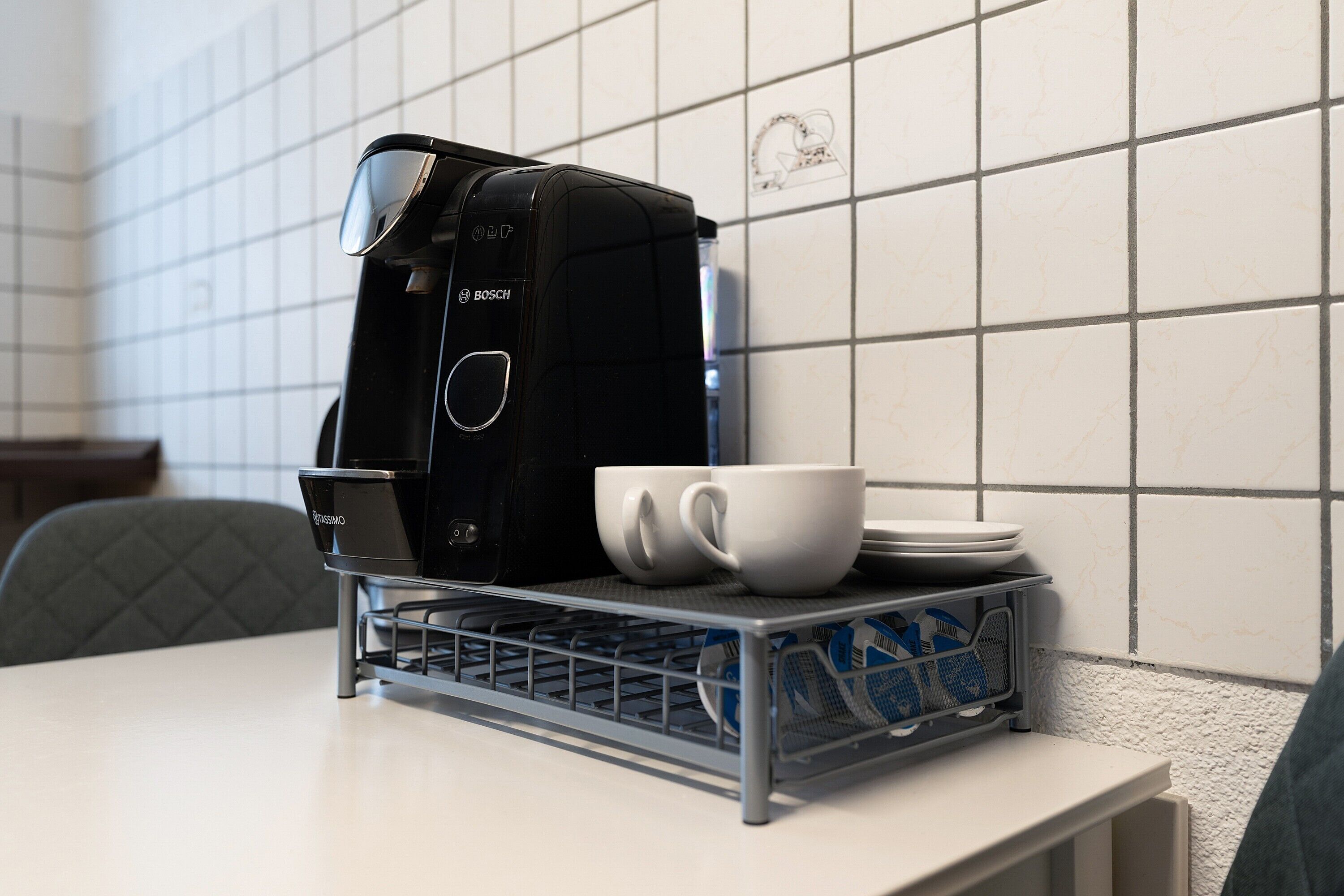 Una cafetera sobre una mesa con una bandeja que contiene tazas y platos, y una pared de azulejos al fondo.