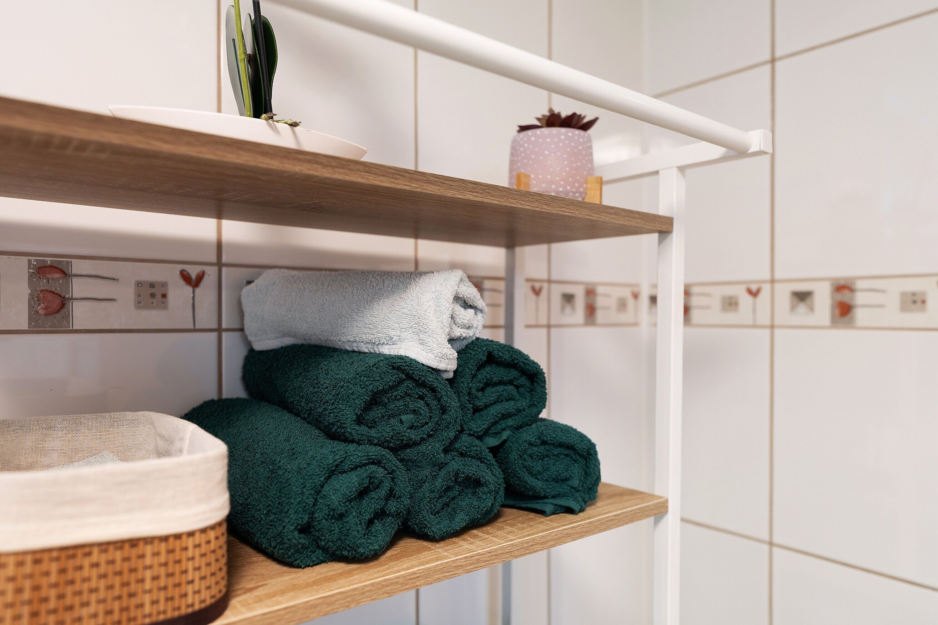 Een badkamer met een houten plank waarop opgerolde handdoeken, een witte container en een plant in pot staan.