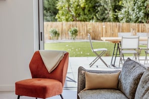 A modern living room with a red armchair and a grey sofa, both with throw pillows. A glass door leads to a patio with a wooden fence and a table set for dining.