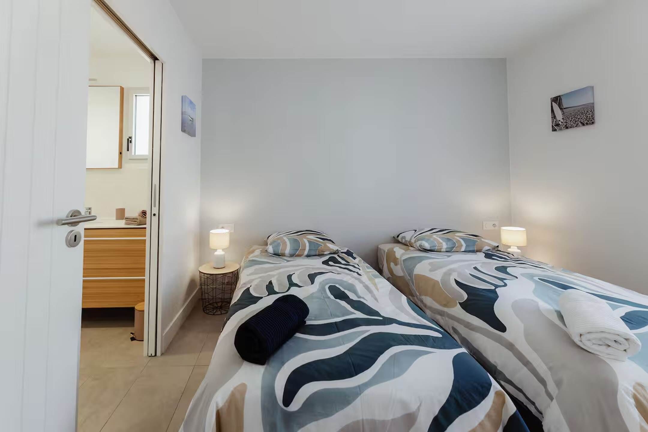A hotel room with two beds, each with a patterned bedspread and pillows, a bedside table with a lamp, and a wooden dresser in the background.