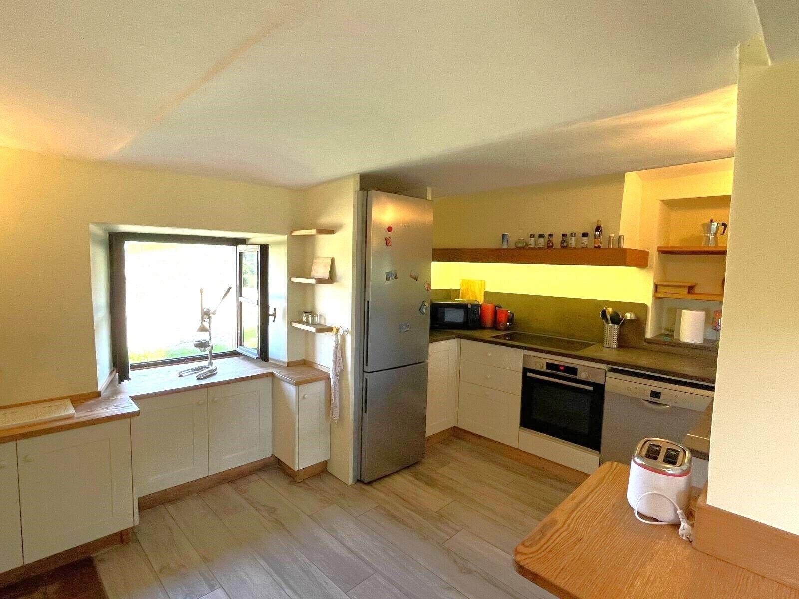 Uma cozinha compacta com geladeira, micro-ondas, forno e torradeira. Há uma janela com vista para o exterior e piso de madeira.
