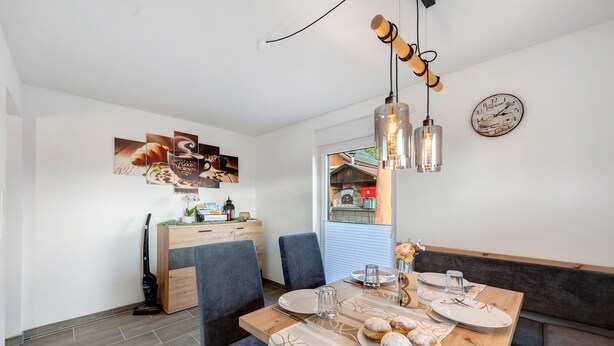 A dining area with a wooden table set for four, a bench, and a view of a red building through a window. - Ferienwohnung Schäfer – Nature, Comfort & Relaxation in the Black Forest (Freudenstadt)