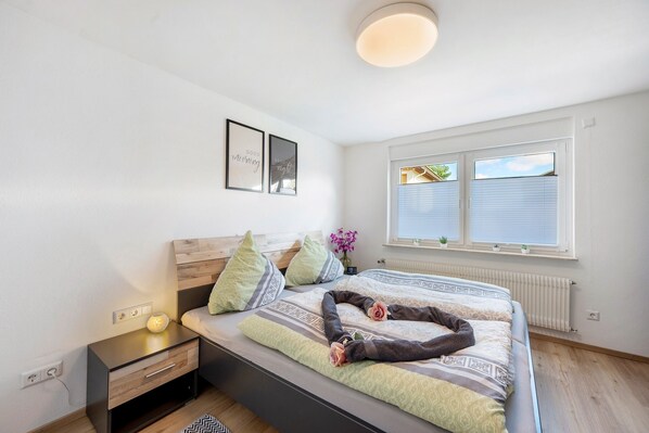 A bedroom with a bed, bedside table, wall art, window with blinds, and a small plant on the windowsill.