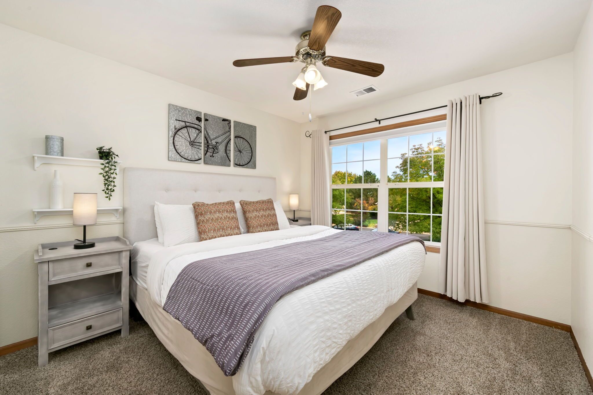 Un dormitorio con cama, mesitas de noche, ventilador de techo, ventana con cortinas y decoración en las paredes.